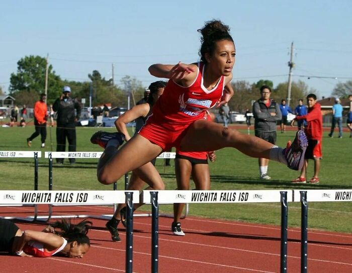 Athlete mid-air jumping hurdle during race, unexpectedly captured with competitor falling on the track behind her.