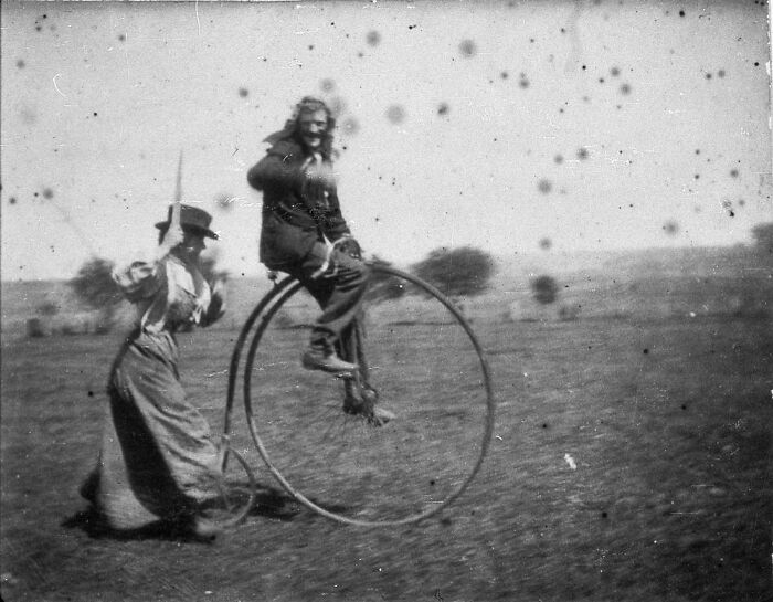 A candid old photo of a man on a penny-farthing bicycle being pushed by another man, revealing what the world looked like generations ago.