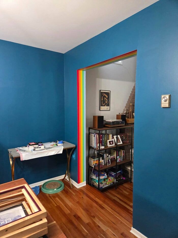 Room with bold blue walls and a rainbow-trimmed doorway featuring shelves filled with books and decor, embracing more is more home style