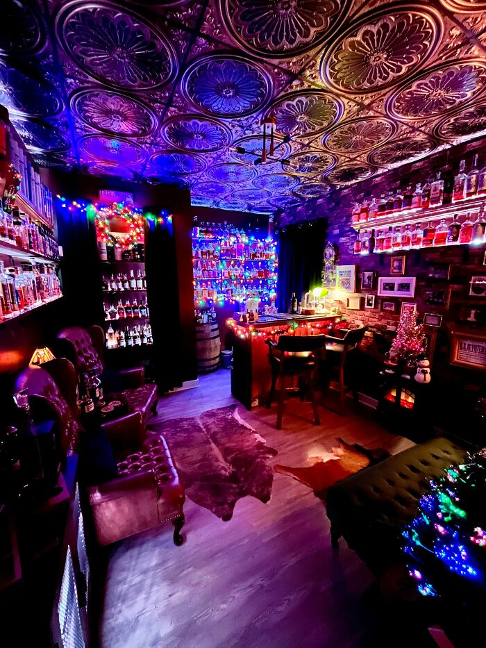 Cozy home bar decked out with festive lights, shelves filled with bottles, leather chairs, and detailed patterned ceiling.