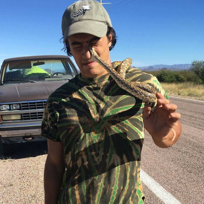 Man wearing camo shirt and cap has snake touching his face, an unexpected perfect photo moment on roadside.