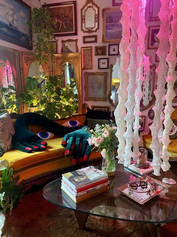 Eclectic living room embracing more is more decor with large plush hands, plants, flowers, mirrors, and layered art on walls.