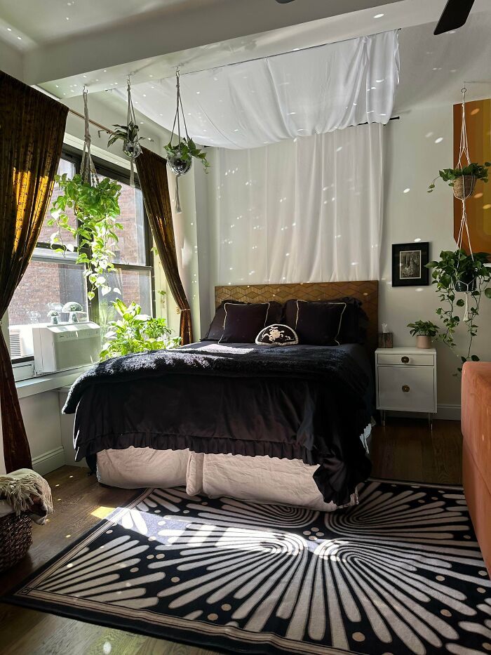 Cozy bedroom embracing more is more style with hanging plants, dark bedding, patterned rug, and layered window treatments.