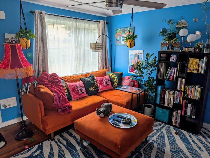 Colorful living room embracing more is more style with bright orange sofa, patterned pillows, eclectic decor, and abundant greenery.