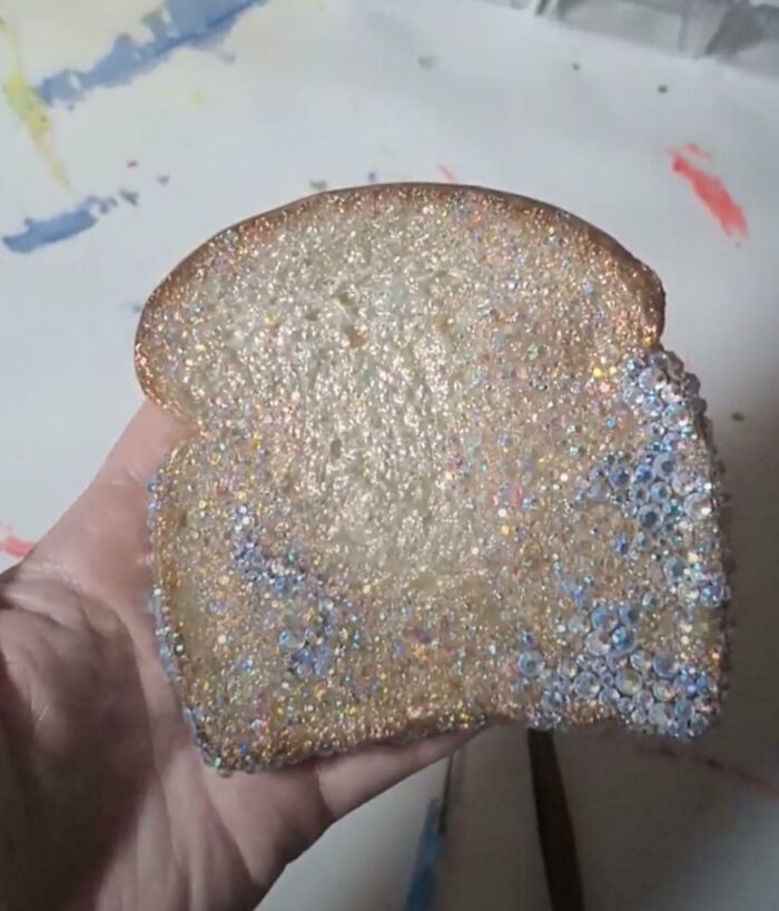 Hand holding a DIY project slice of bread covered in glitter and crystals, showing a deeply cursed craft result.