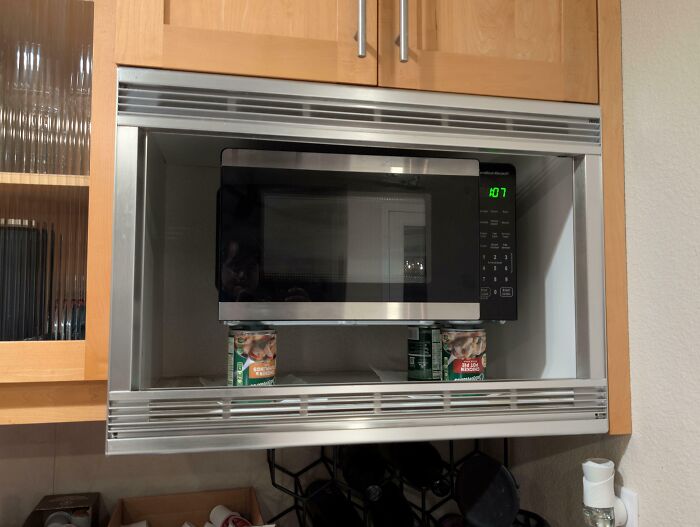 Microwave elevated by cans inside a kitchen cabinet showing a deeply cursed DIY project fail.
