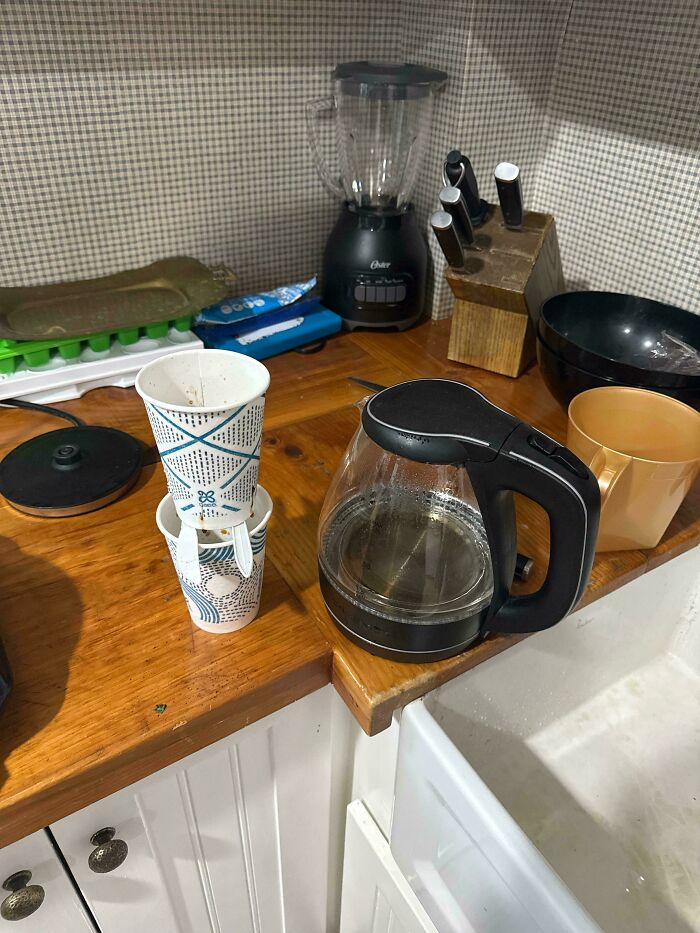 DIY project fail with stacked paper cups on kitchen counter beside coffee pot and utensils, showcasing a deeply cursed setup.