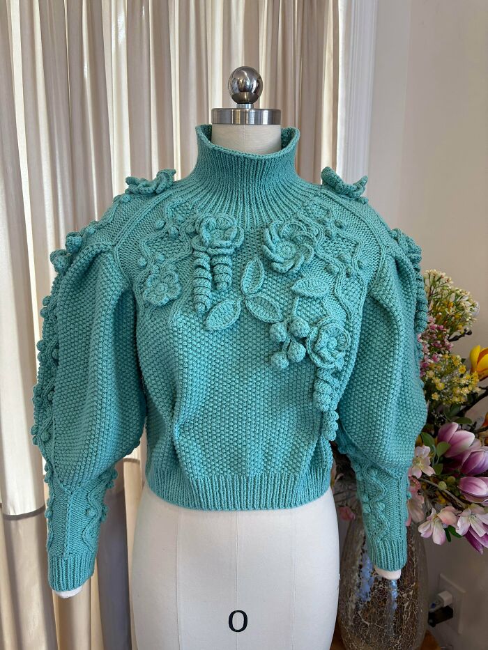 A stunning teal crocheted sweater on a mannequin, featuring intricate 3D floral designs and puffy sleeves. People crocheted something awesome here!