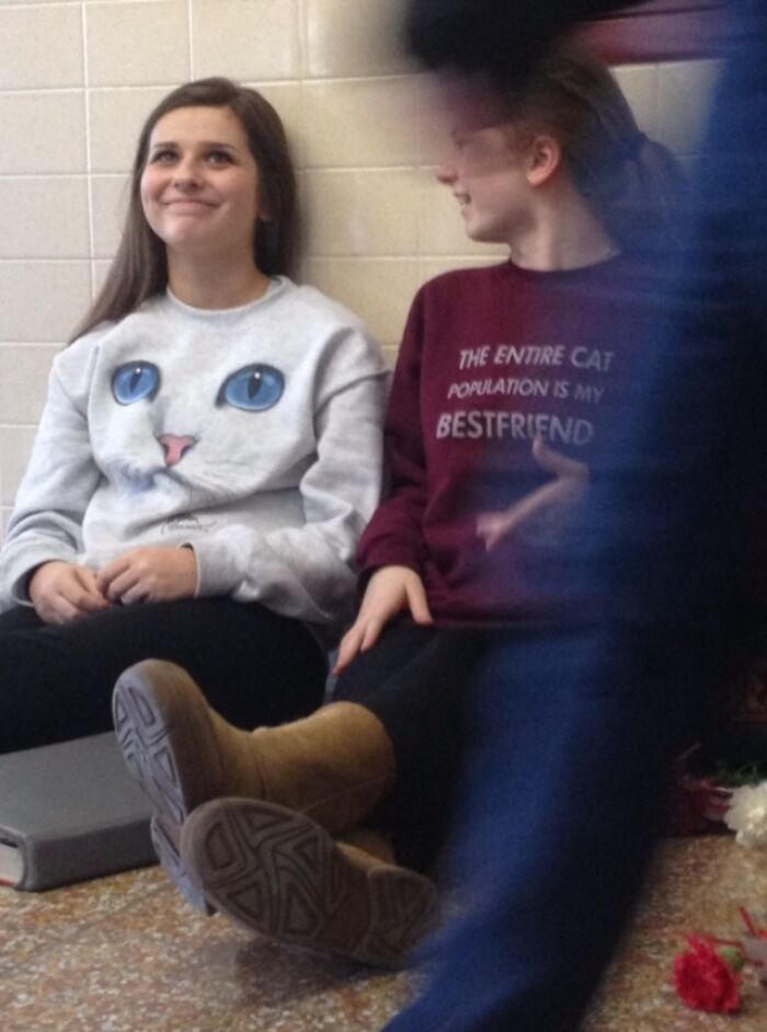 Two young women sitting and smiling, one wearing a funny cat face sweatshirt, capturing a funny photo moment.