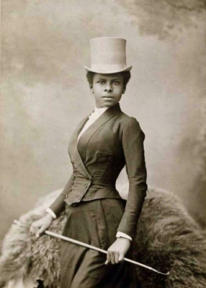 Victorian era photo of a confident woman in period attire with a top hat and riding crop, showcasing fashion of the time.