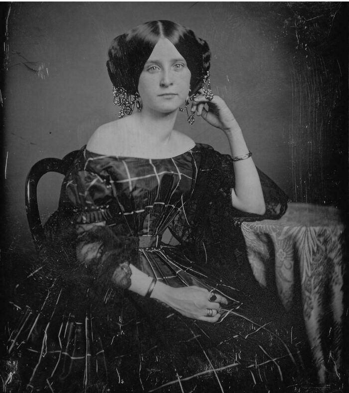 Victorian era woman in period dress and detailed hairstyle seated next to a table with patterned cloth, vintage photo.