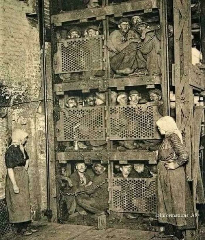 Victorian era photo showing miners crowded inside metal cages with women standing nearby at an industrial site.
