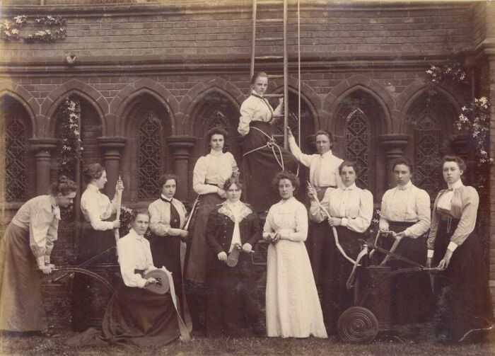 Group of women in Victorian era clothing posing outdoors by a brick building with arched windows in a vintage Victorian era photo.