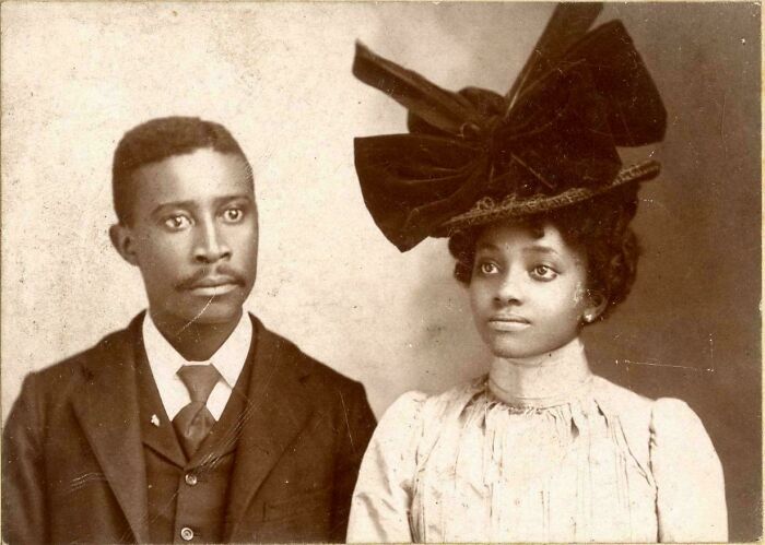 Victorian era photo of a man in a suit and a woman wearing a large hat and period dress in a vintage portrait.