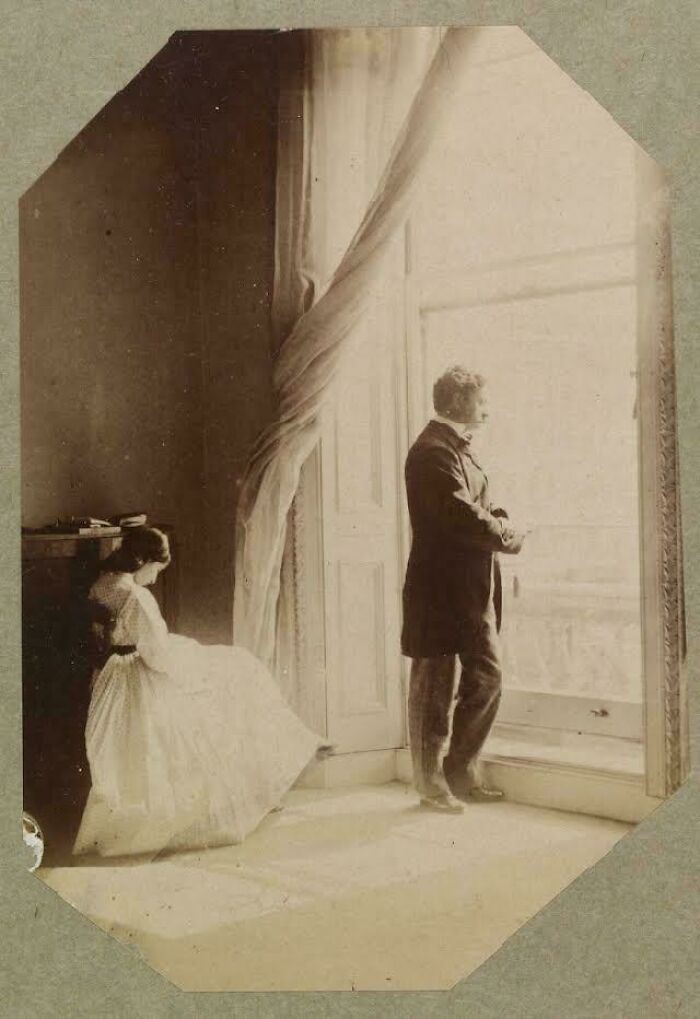 Victorian era photo of a woman sitting near a man standing by a large window with flowing curtains inside a room.
