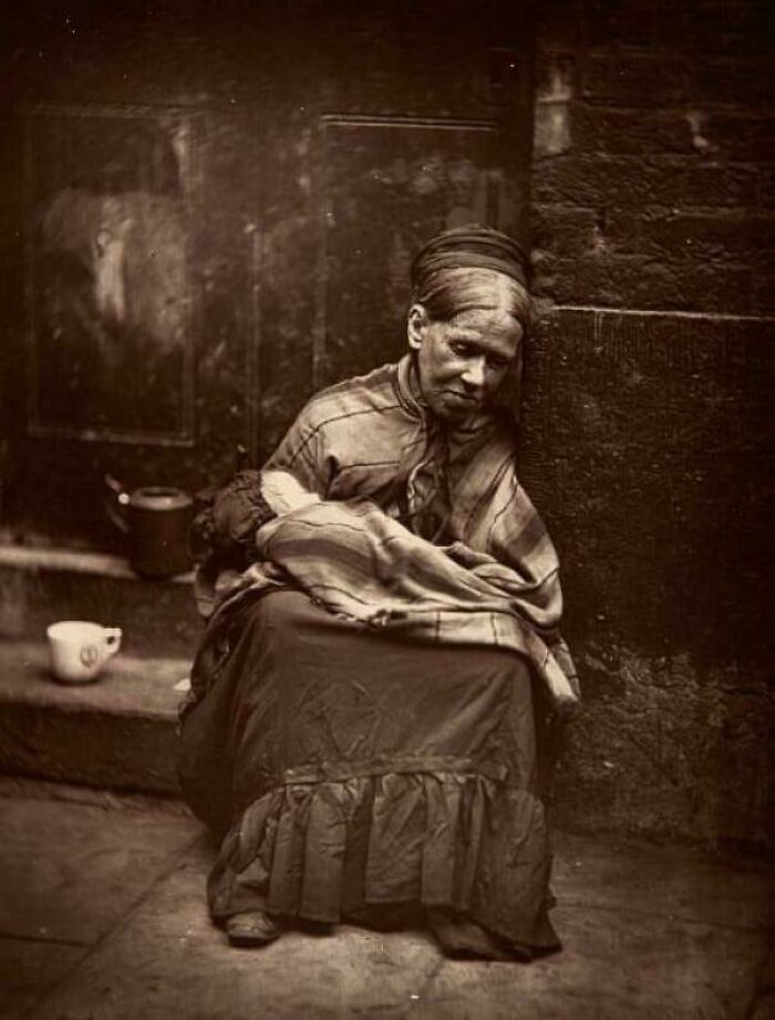 Victorian Era photo of a woman sitting on a street corner holding a wrapped bundle, wearing period clothing.