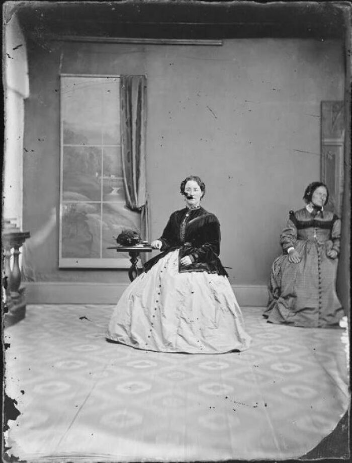 Two women dressed in Victorian era clothing seated in a sparsely decorated room with patterned flooring.