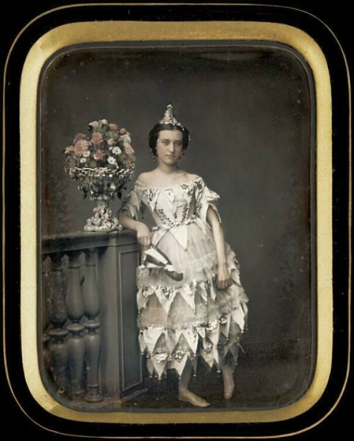 Victorian era photo of a woman in an elaborate dress and tiara, standing beside a floral arrangement on a balcony railing.