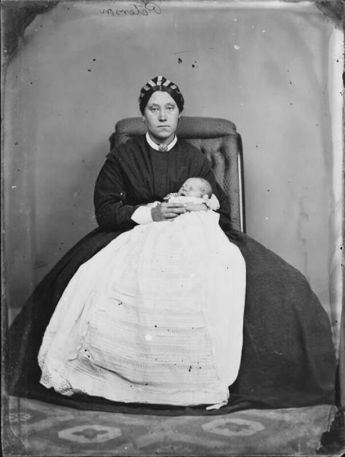 Victorian era woman seated in a chair holding a baby wrapped in a white blanket in a historic black and white photo.