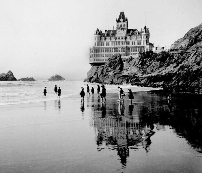 Children in Victorian Era photos playing on a beach with a large cliffside building reflected in the wet sand.