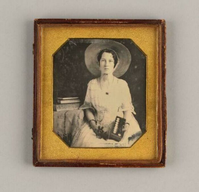 Victorian era photo of a seated woman in period dress and hat, holding an object, framed in a vintage case.