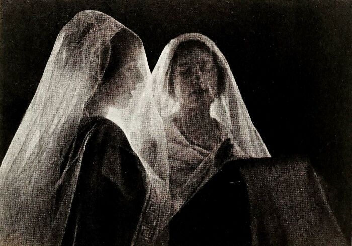 Two Victorian era women wearing veils stand closely together reading from a book in a dimly lit setting.