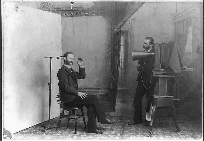 Victorian era photo of a man seated for an early photography session with a photographer using vintage equipment.