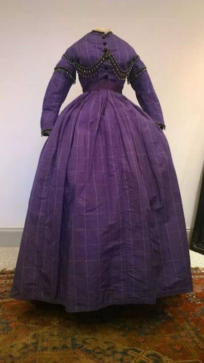 Victorian era dress in purple fabric with full skirt and decorative trim, showcasing period fashion details.