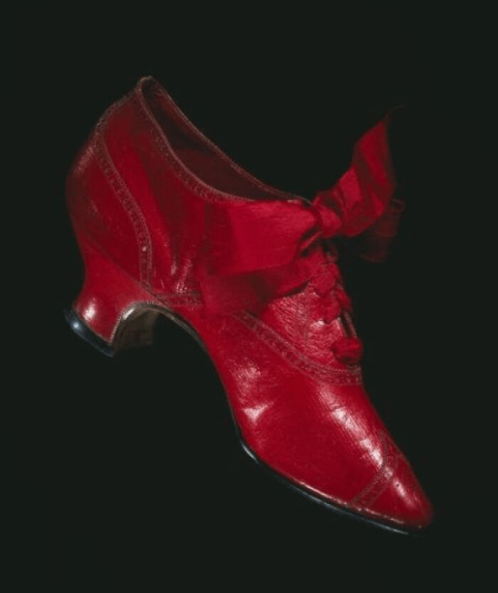 Victorian era red leather shoe with a bow and low heel, showcasing period fashion details against a black background.