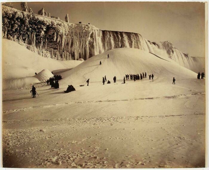 Victorian era photo showing a large frozen waterfall with people exploring snowy ice formations near it.