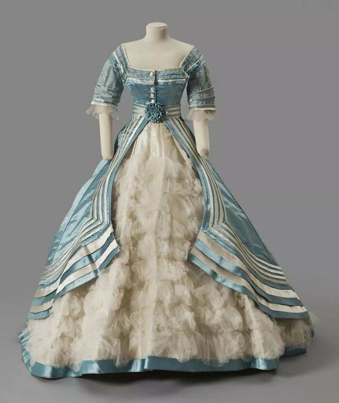 Victorian era dress in blue and white silk with intricate detailing on a mannequin against a plain background.