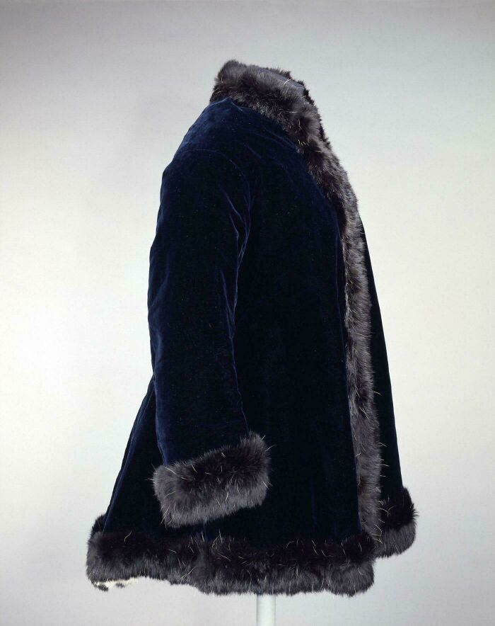 Victorian era coat in dark velvet with fur trim on cuffs, collar, and hem, showcasing period fashion details.