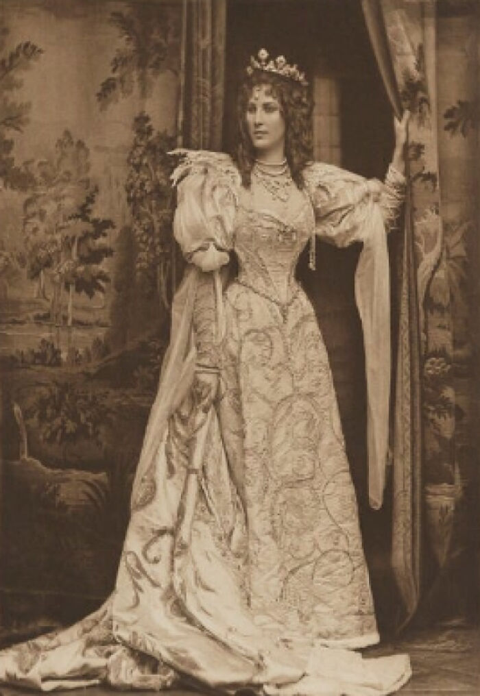 Victorian Era photo of a woman in an ornate, long gown and crown, standing by a curtain in a detailed indoor setting.