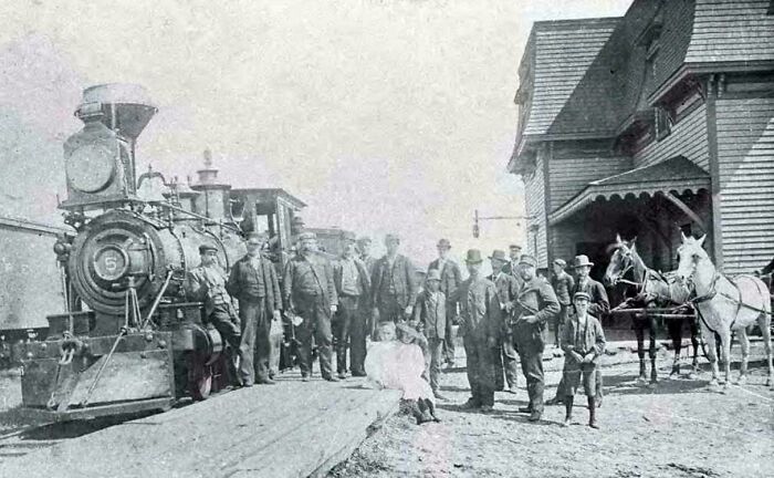 Victorian era photo of people, horses, and a steam locomotive at a rural train station, showcasing period transportation.