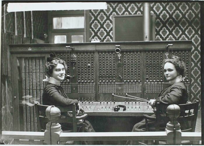Two women operating a Victorian era telephone switchboard in a patterned room from the historical period.
