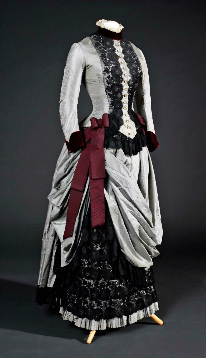 Victorian era dress with black lace and burgundy bows displayed on a mannequin against a dark background.