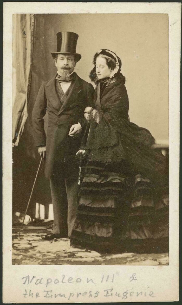 Victorian era photo of a man in top hat and a woman in elaborate dress posing indoors with rich period clothing details.