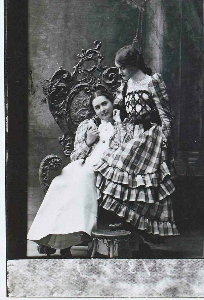 Two women in Victorian era dresses posing by an ornate carved wooden chair in a vintage photo.
