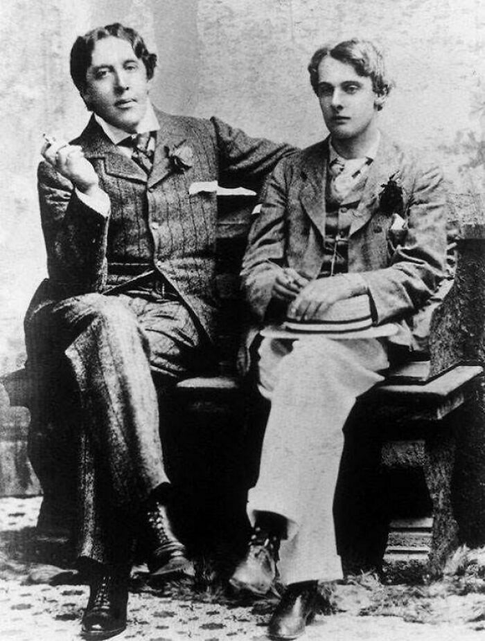 Two men in Victorian Era photos dressed in period suits, seated on a bench in a vintage black and white image.