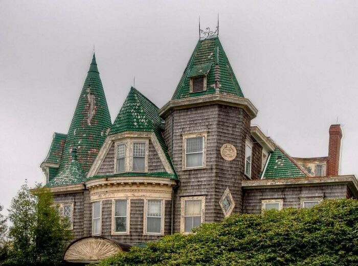 Victorian era house with worn green rooftops and wooden siding surrounded by bushes under an overcast sky.