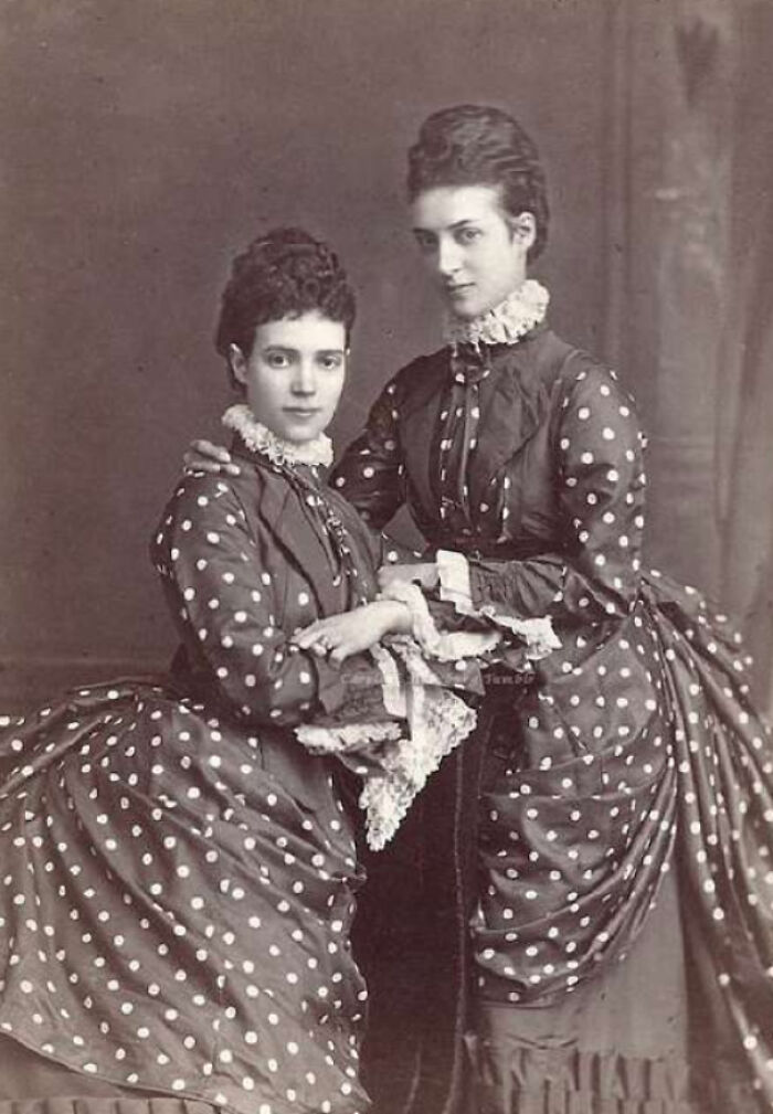 Two Victorian era women in polka dot dresses posing for a formal portrait in the late 19th century.