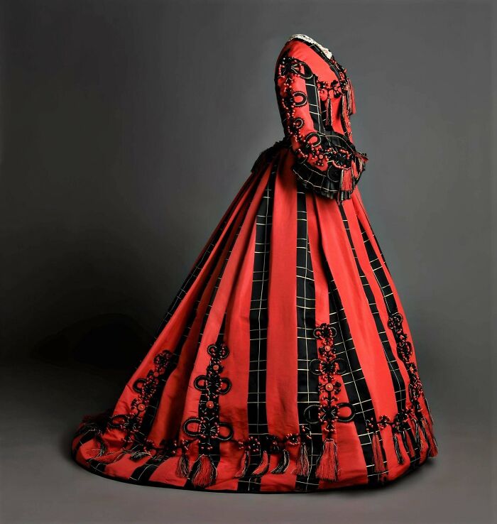 Victorian era dress in red and black with intricate patterns and tassels, showcasing 19th century fashion details.