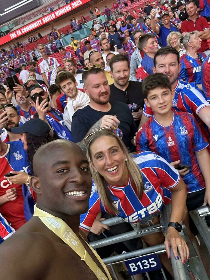 Crowd of sports fans in stadium wearing red and blue jerseys, a selfie with a smiling shirtless man and group behind him.