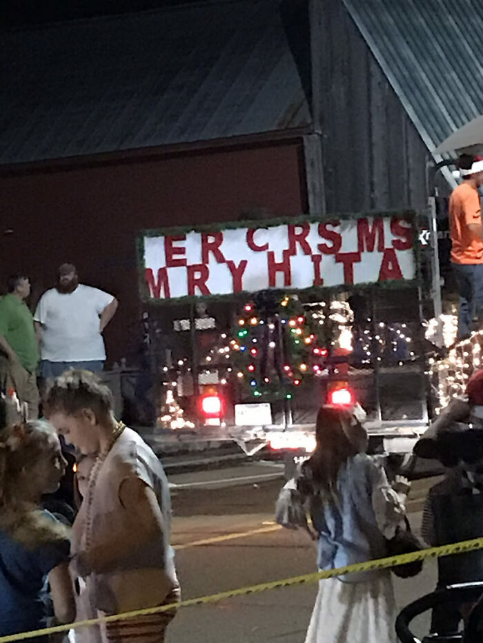 Sign with Christmas message gone wrong, featuring blurred letters and decorated with holiday lights at night event.