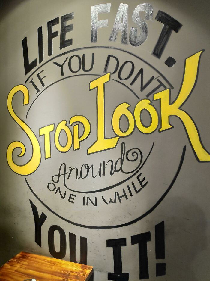 Wall sign with comedic gold message about life going fast, featuring bright yellow and black hand-painted lettering on gray background.