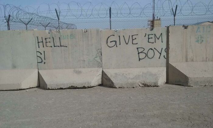 Concrete barriers with graffiti text saying "HELL" and "GIVE 'EM BOYS" under barbed wire fence, a sign fail turned comedic gold.