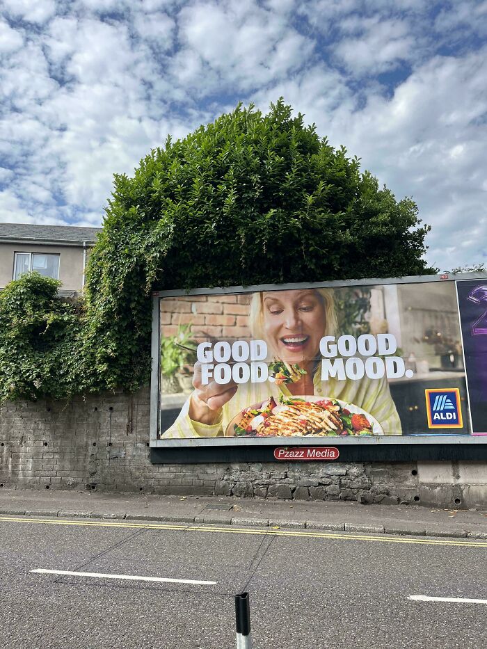 Billboard sign with a woman eating food partially obscured by overgrown greenery, a comedic sign fail example.
