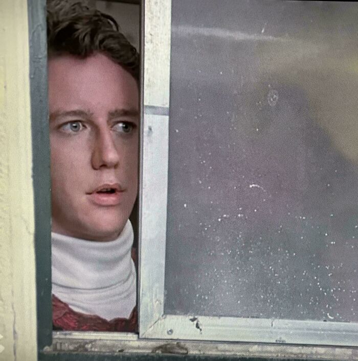 Young man with light brown hair and a white turtleneck peeking out from behind a window, an image that takes you way back.