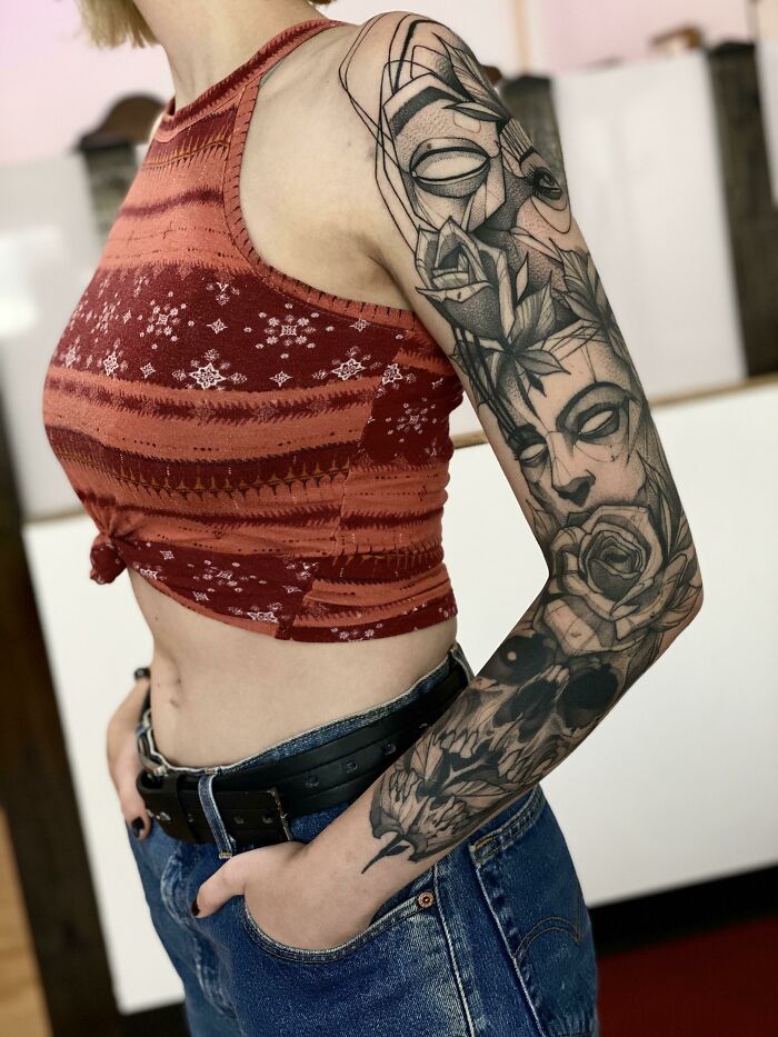 Woman wearing a patterned crop top showing a detailed black and grey cool tattoo sleeve with faces and floral elements.