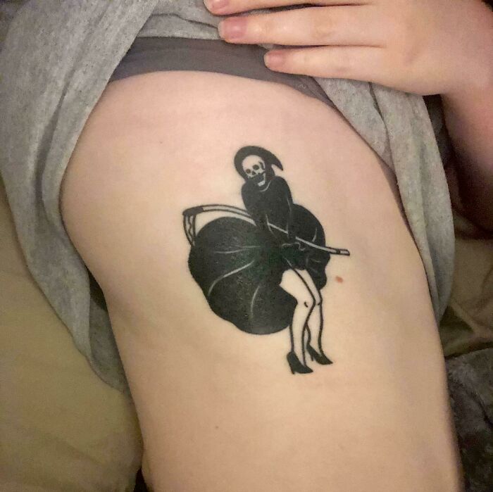 Black ink tattoo of a skeleton figure wearing a dress and high heels holding a scythe on a person's side.